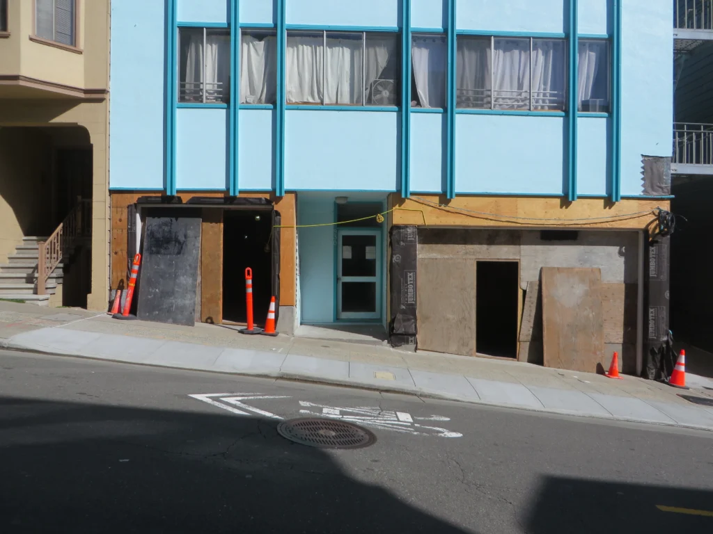 A blue building under construction on a sloped street, with plywood barriers, construction cones, and an open doorway visible at street level.