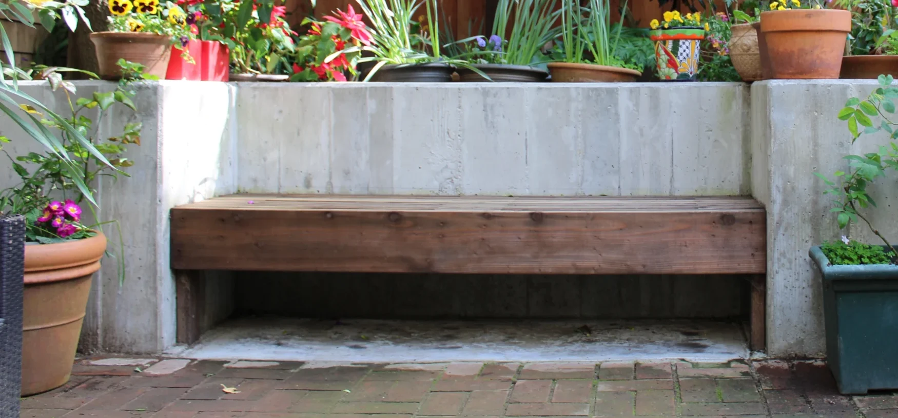 A wooden bench is set against a concrete planter filled with various flowering plants and surrounded by potted plants in a fenced backyard garden.