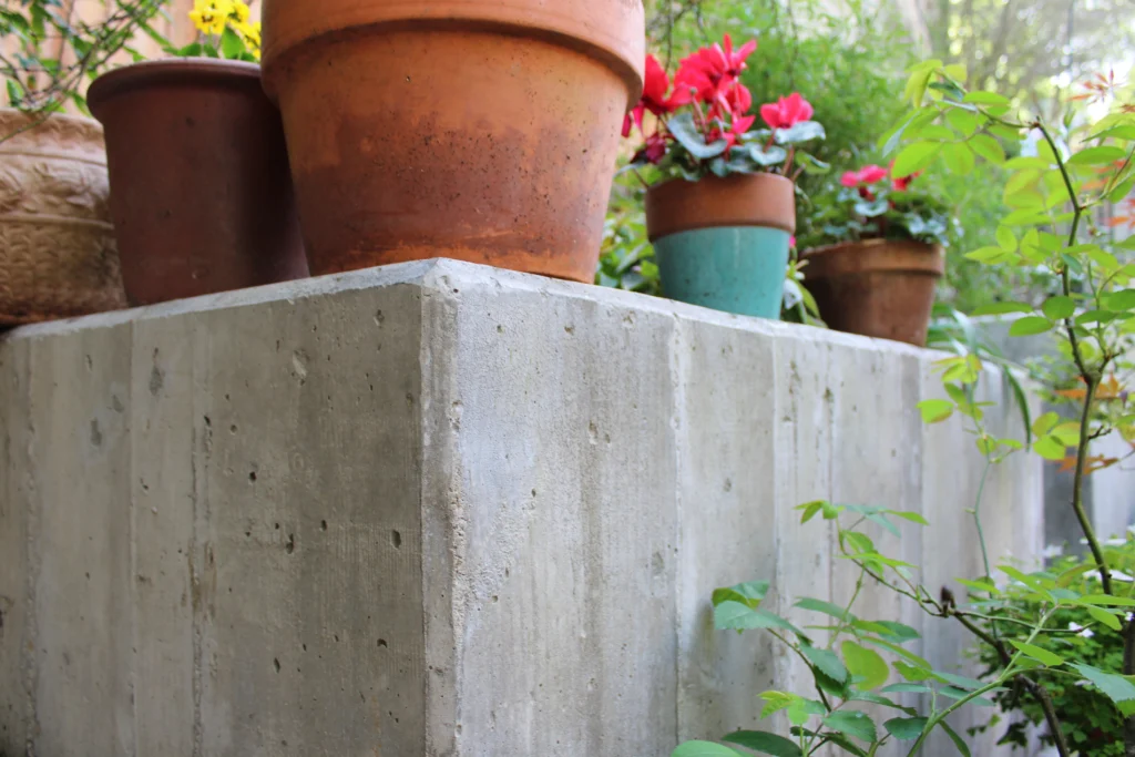 Several potted plants, including red flowers, are arranged on top of a concrete block outdoors with green foliage nearby.