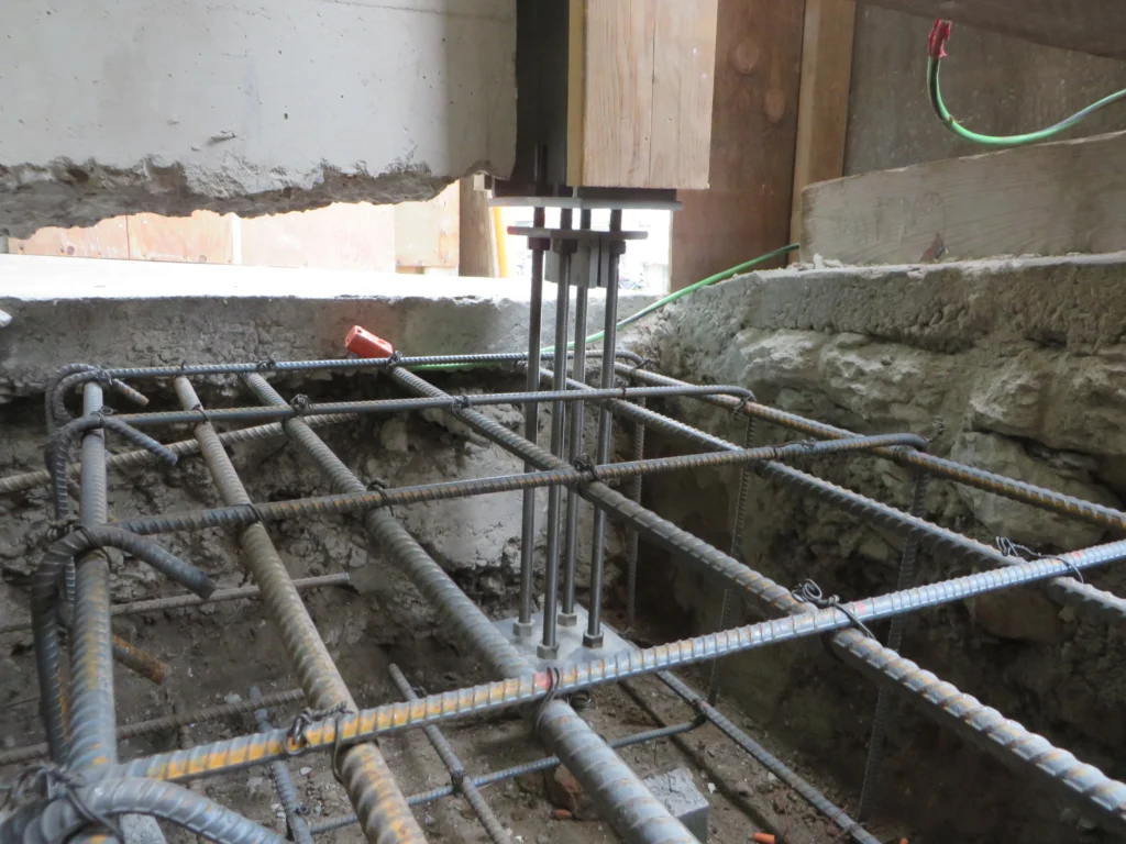 Steel reinforcement bars and anchor bolts embedded in a concrete foundation, with exposed rebar grid and surrounding unfinished construction materials.