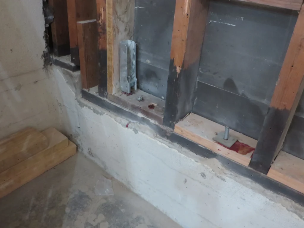 Exposed wall framing and anchor bolts in a basement, with some wood, concrete, and metal hardware visible, and minor red staining on the sill plate.