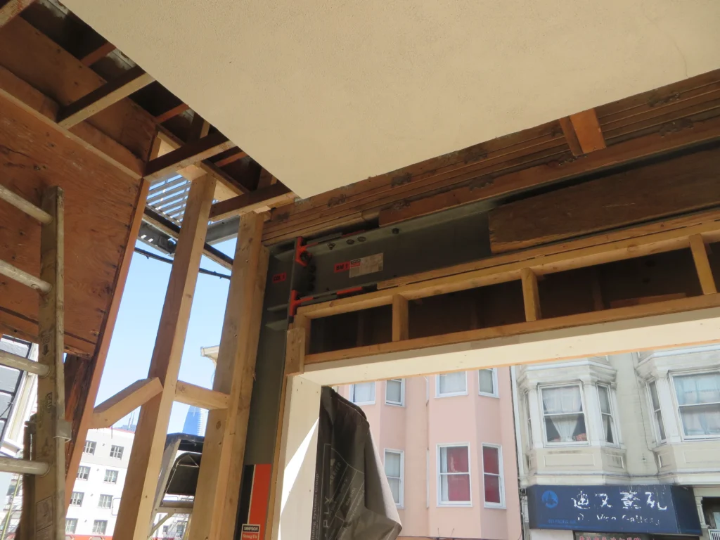 Interior view of a building under construction, showing exposed wooden framing, steel beam, support braces, and part of an open doorway leading to a city street with neighboring buildings.