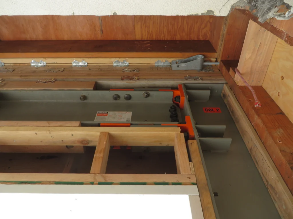 A close-up of a structural steel beam with bolts, brackets, and wood framing in a partially constructed building. Labels and a diagram are visible on the beam.