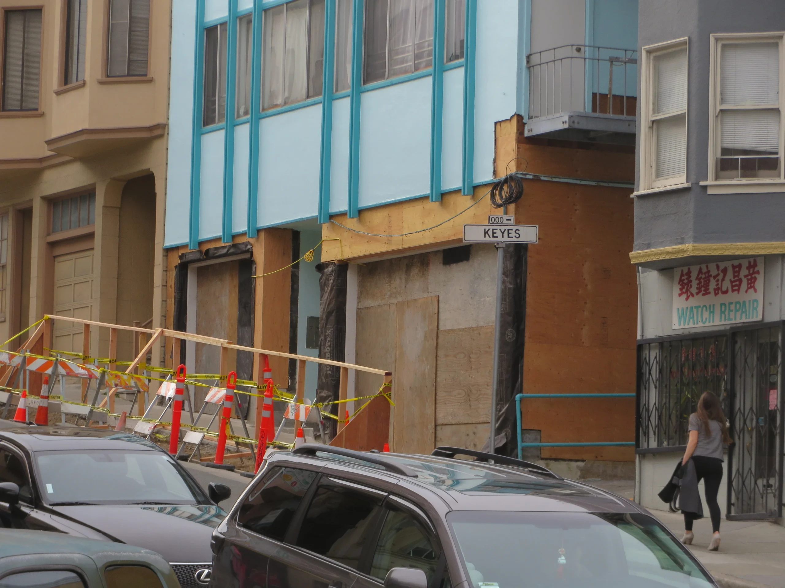 A building under construction with boarded-up storefronts and orange cones, located at the corner of Keyes street. A woman walks past a watch repair shop.