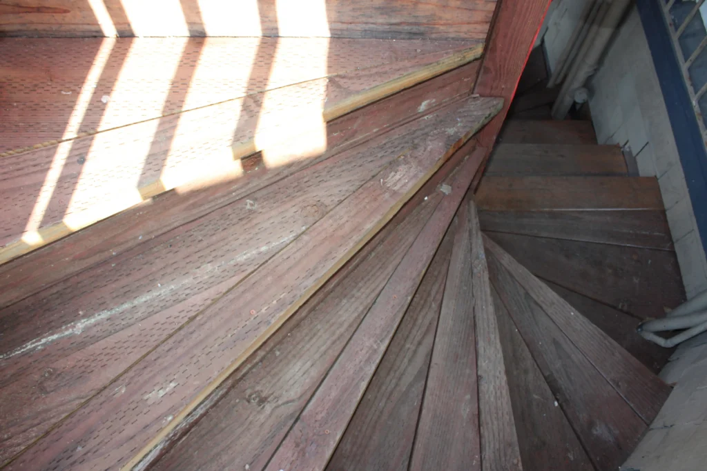 A top-down view of a spiral wooden staircase with worn, narrow steps and sunlight casting shadows on the upper landing.