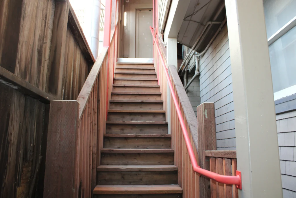 A wooden staircase with red handrails leads up to a closed beige door, situated between two buildings with wood siding.