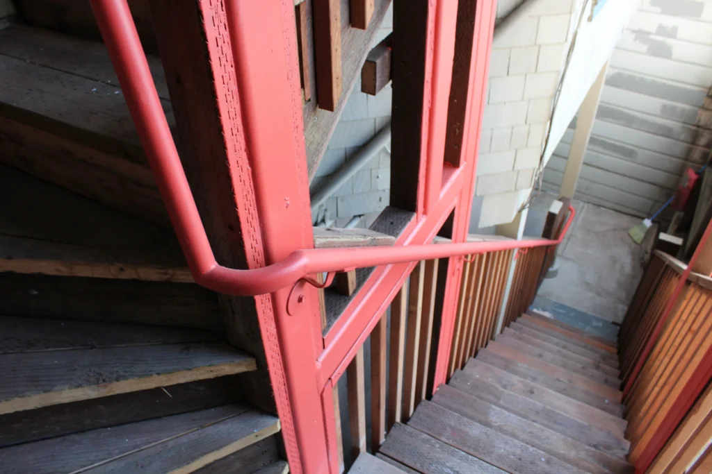 A red metal handrail runs alongside a wooden staircase with wooden and concrete walls in the background.