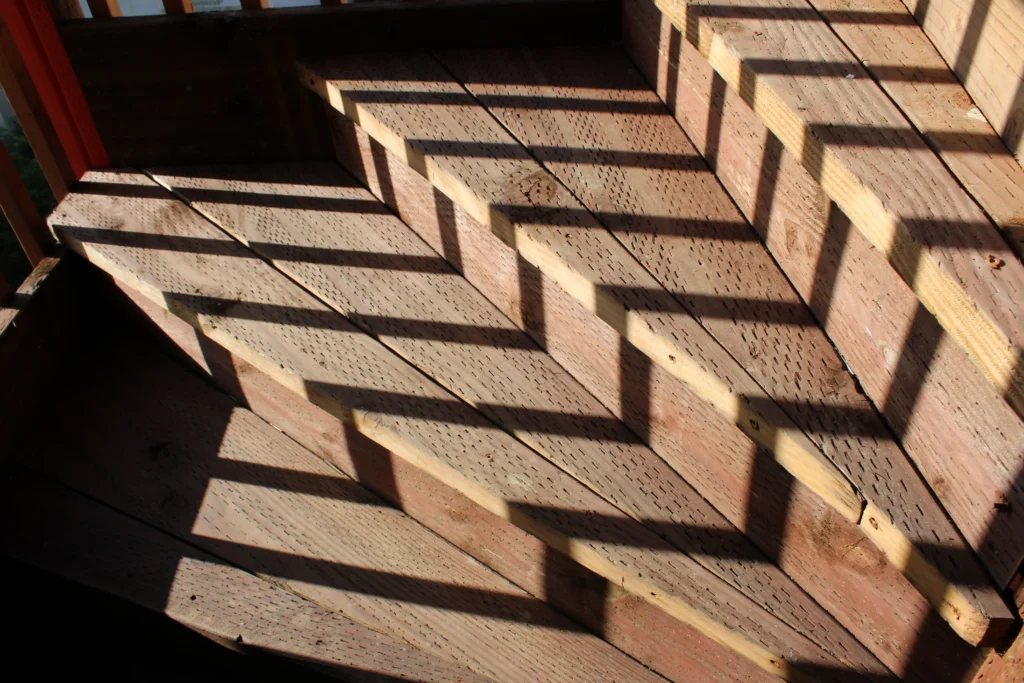 Wooden stairs with strong diagonal shadows cast by sunlight, creating a repeating pattern of light and dark across the steps.