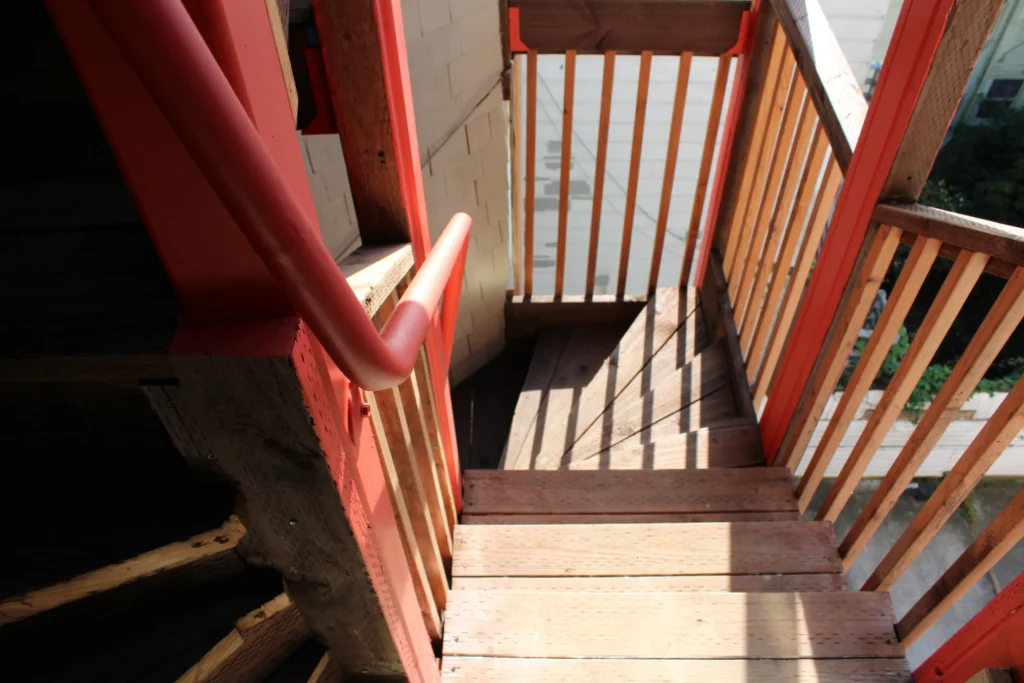 Wooden outdoor staircase with red railings, leading down to a small landing. Sunlight casts shadows across the steps and railing.